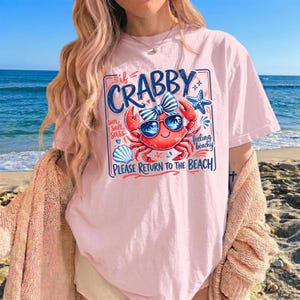 May include: A light pink t-shirt with a graphic of a crab wearing sunglasses and a bow tie. The text on the shirt reads "If Crabby, Please Return to the Beach." The design includes a seashell, starfish, and the words "Sun, Salt, Sass, Feeling Beachy."
