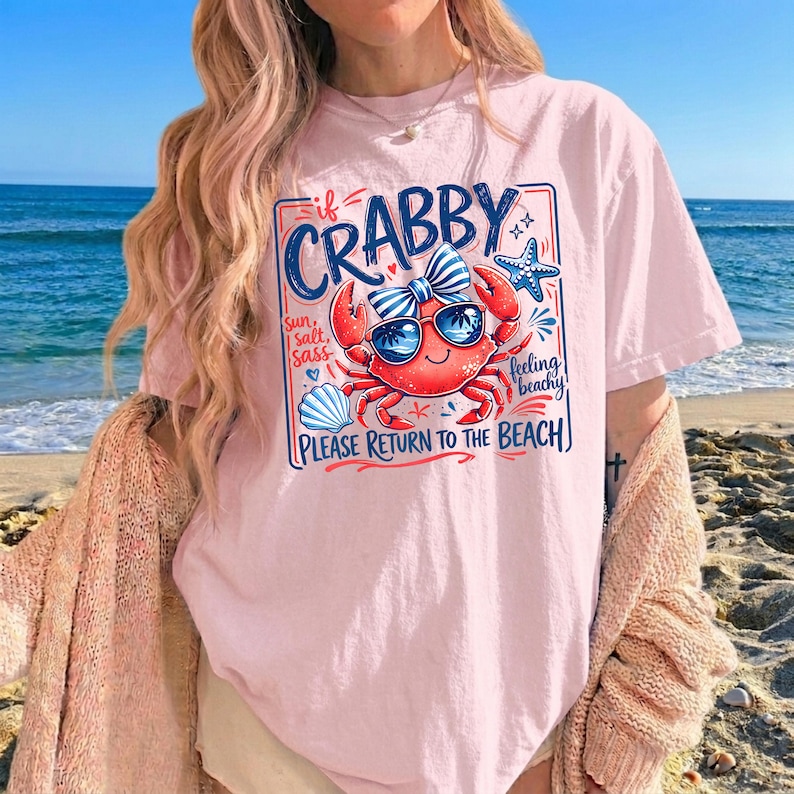 May include: A light pink t-shirt with a graphic of a crab wearing sunglasses and a bow tie. The text on the shirt reads "If Crabby, Please Return to the Beach." The design includes a seashell, starfish, and the words "Sun, Salt, Sass, Feeling Beachy."