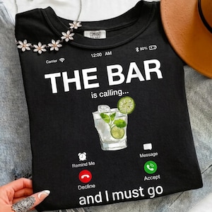 Könnte beinhalten: Schwarzes T-Shirt mit dem Text "THE BAR is calling... and I must go." Eine Grafik eines Cocktails mit Limettenscheiben ist abgebildet. Das Design enthält Telefon-ähnliche Symbole zum Ablehnen, Annehmen und für Erinnerungen.