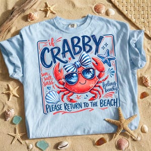 Pode incluir: Camiseta azul clara com um desenho de um caranguejo vermelho usando óculos de sol e uma gravata borboleta. O texto na camisa diz "If Crabby, Please Return to the Beach". O design inclui uma estrela do mar e uma concha.