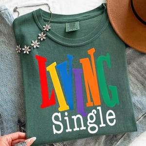 K&ouml;nnte beinhalten: Ein dunkelgr&uuml;nes T-Shirt mit den Worten "LIVING SINGLE" in bunten Blockbuchstaben. Die Buchstaben sind rot, gelb, blau, lila, orange und gr&uuml;n. Eine silberne Halskette mit Blumenakzenten liegt auf dem Shirt.