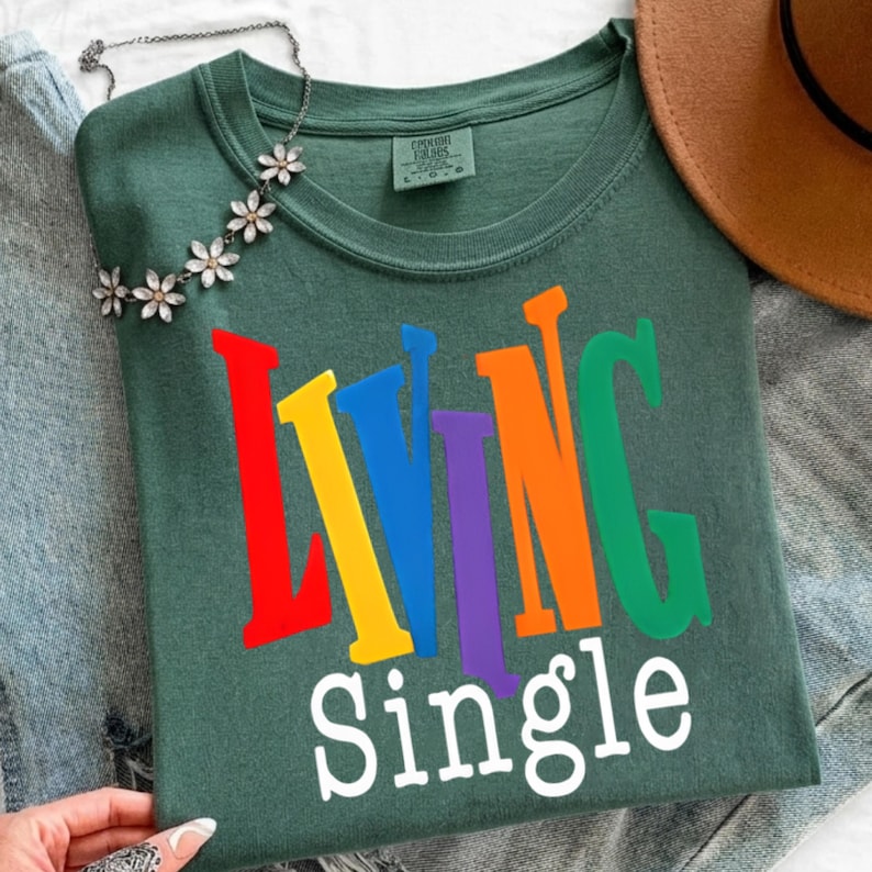 K&ouml;nnte beinhalten: Ein dunkelgr&uuml;nes T-Shirt mit den Worten "LIVING SINGLE" in bunten Blockbuchstaben. Die Buchstaben sind rot, gelb, blau, lila, orange und gr&uuml;n. Eine silberne Halskette mit Blumenakzenten liegt auf dem Shirt.