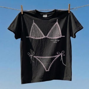 May include: Black t-shirt with a white line drawing of a bikini top and bottom. The words "Palm Springs 2026" are written in cursive below the bikini top. The shirt is hanging on a clothesline.