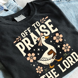 May include: A black t-shirt featuring a graphic of a white goose wearing a pink bow and holding a brown book. The text reads "OFF TO PRAISE THE LORD" in cream-colored letters, surrounded by small pink flowers, green leaves, and white stars.