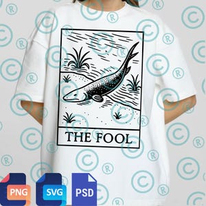 May include: White t-shirt featuring a black and white illustration of a fish with the text "THE FOOL" in a black rectangle. The design includes aquatic plants and lines. The shirt is a casual, oversized fit.