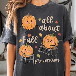 May include: A charcoal gray t-shirt featuring a graphic of three cartoon pumpkins. One pumpkin uses a cane, one uses a walker, and one sits in a wheelchair. The text reads "all about Fall prevention" in a light tan script font with small autumn leaves.