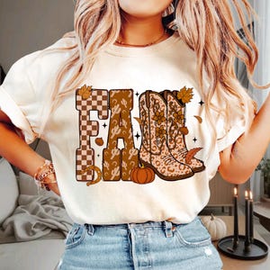 May include: A cream-colored t-shirt featuring a graphic print of the word FALL. The letters are decorated with a brown and white checkerboard pattern, a cactus print, and a pair of leopard-print cowboy boots with floral accents and autumn leaves.