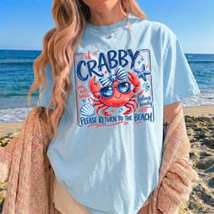 May include: Light blue t-shirt featuring a cartoon crab wearing sunglasses and a bow, with the text "If Crabby, Please Return to the Beach." The design includes a starfish and seashell. The shirt is worn on a beach.