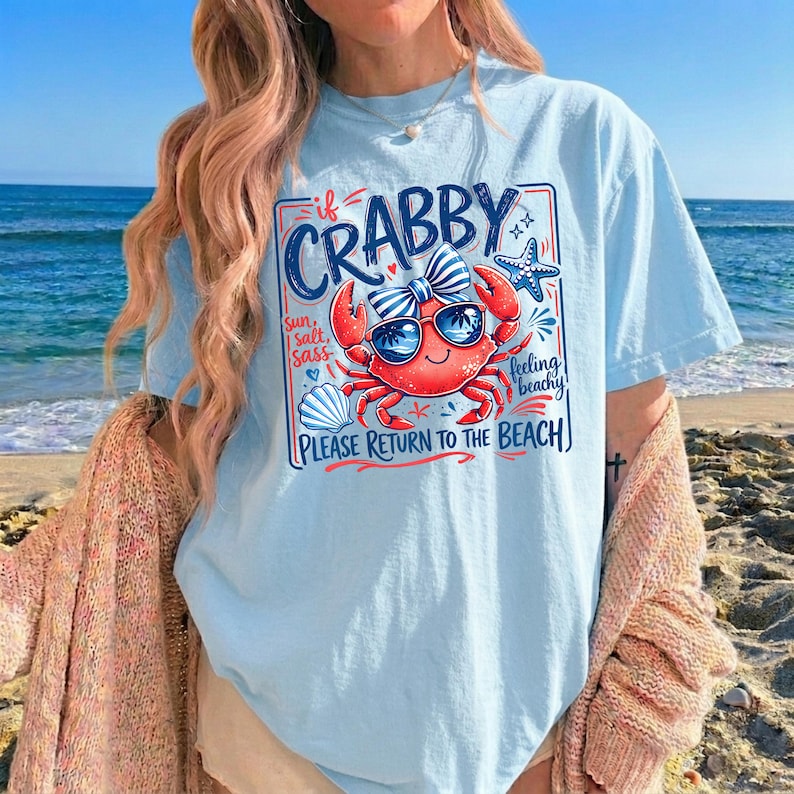 May include: Light blue t-shirt featuring a cartoon crab wearing sunglasses and a bow, with the text "If Crabby, Please Return to the Beach." The design includes a starfish and seashell. The shirt is worn on a beach.