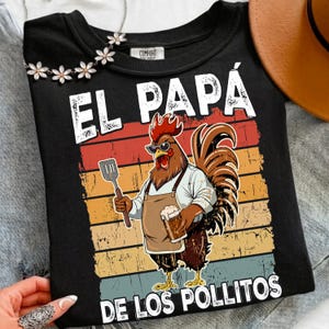 Puede incluir: Una camiseta negra con un gráfico de un gallo con gafas de sol y un delantal, sosteniendo una espátula y un vaso de cerveza. El texto dice EL PAPA DE LOS POLLITOS. El diseño de fondo consiste en rayas horizontales de color naranja y azul.