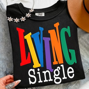 Könnte beinhalten: Schwarzes Sweatshirt mit Rundhalsausschnitt und dem Schriftzug "Living Single" in bunten Buchstaben. Eine Blumenkette und ein brauner Hut sind im Hintergrund zu sehen.