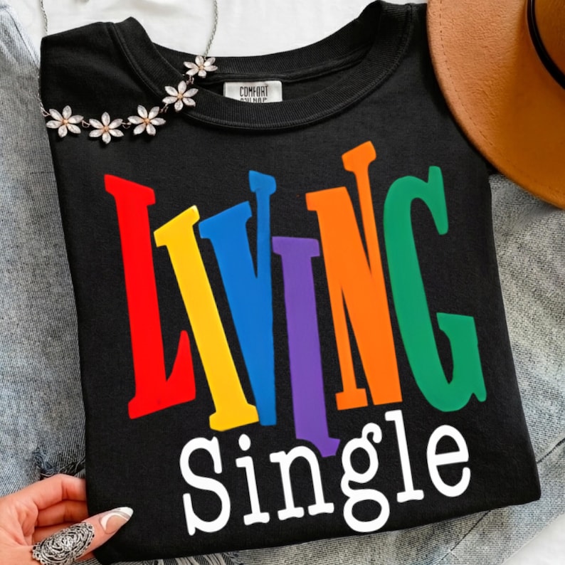 K&ouml;nnte beinhalten: Schwarzes Sweatshirt mit Rundhalsausschnitt und dem Schriftzug "Living Single" in bunten Buchstaben. Eine Blumenkette und ein brauner Hut sind im Hintergrund zu sehen.
