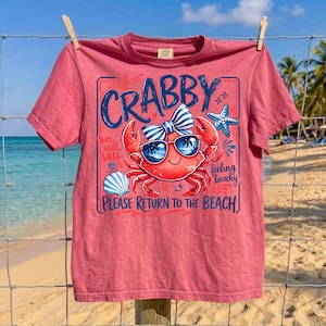 May include: A red t-shirt with a crab graphic wearing sunglasses and a bow tie. The text on the shirt reads "Crabby" and "Please Return to the Beach". The shirt is hanging on a clothesline with a beach background.