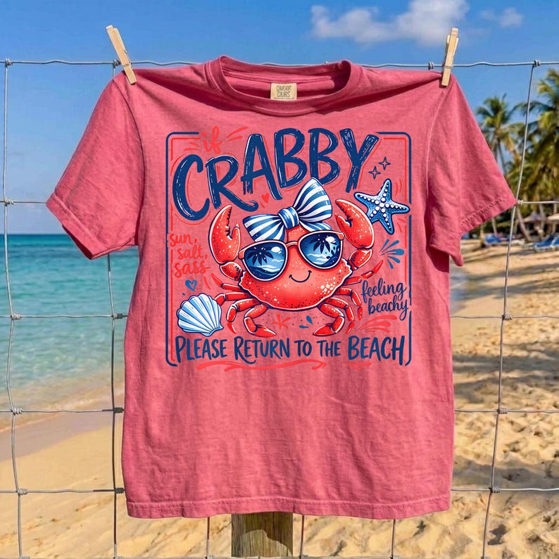 May include: A red t-shirt with a crab graphic wearing sunglasses and a bow tie. The text on the shirt reads "Crabby" and "Please Return to the Beach". The shirt is hanging on a clothesline with a beach background.