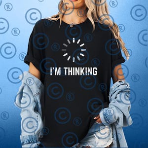 Puede incluir: Camiseta negra con el texto blanco "I'M THINKING" y un símbolo de carga. La camiseta se combina con una chaqueta vaquera y vaqueros azules.