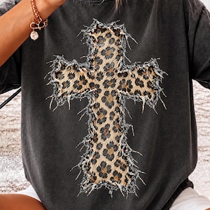 May include: A charcoal gray short-sleeve t-shirt featuring a large, central cross graphic. The cross has a tan and black leopard print pattern and is surrounded by a frayed, distressed-looking border that adds texture to the design.