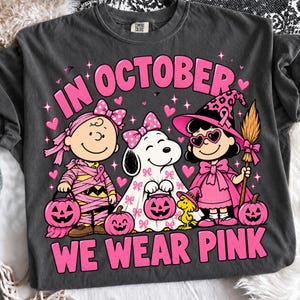 May include: A charcoal grey t-shirt featuring a graphic of three cartoon characters dressed for Halloween. The text reads IN OCTOBER WE WEAR PINK. The characters are wrapped in pink bandages, a ghost sheet with pink bows, and a witch costume with pink accents.