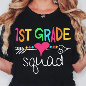 May include: Black t-shirt with the words "1st Grade Squad" in colorful, playful lettering. A white arrow with a pink heart and colorful dots is centered below the text. The shirt is a fun, back-to-school design.
