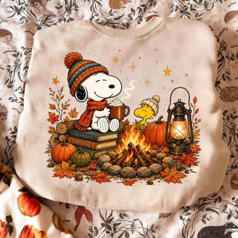May include: A cream-colored sweatshirt featuring a graphic of Snoopy wearing a striped beanie and scarf, sitting on a stack of books labeled Cozy Nights. Beside Snoopy is Woodstock, a campfire, pumpkins, and a lit lantern amidst autumn leaves.
