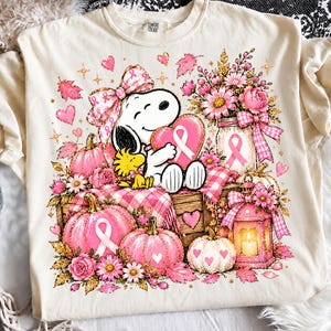 May include: A cream-colored t-shirt featuring a graphic of Snoopy and Woodstock surrounded by pink pumpkins, flowers, a lantern, and hearts. The design includes breast cancer awareness ribbons, a large pink bow, and a plaid blanket pattern.