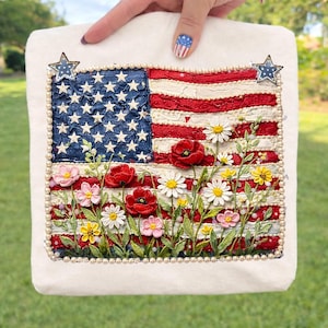以下が含まれることがあります： 白い正方形のバッグ。刺繍されたアメリカ国旗のデザインで、赤と白のストライプ、青い星、花のボーダーが特徴です。バッグは真珠のようなビーズと星型の装飾で飾られ、愛国的なテーマを連想させます。