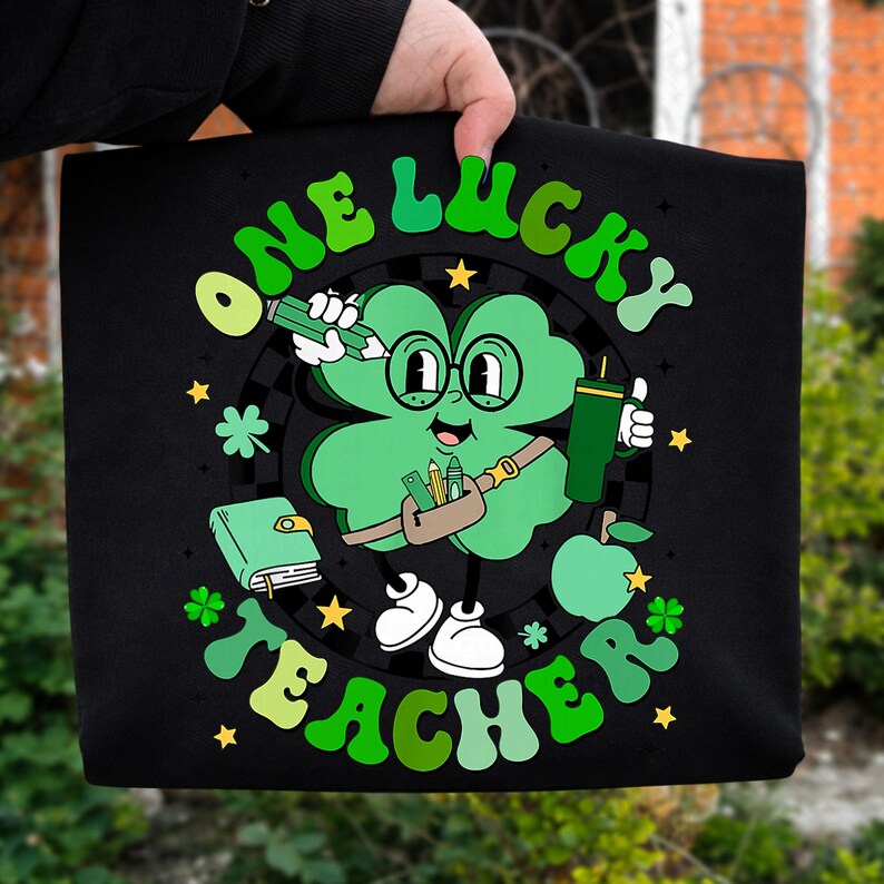 May include: Black tote bag with a green and white graphic of a four-leaf clover character wearing glasses and holding a pencil and a tumbler. The text "ONE LUCKY TEACHER" is in green, retro-style lettering.