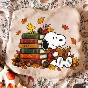 May include: A beige crewneck sweatshirt featuring a graphic print of Snoopy wearing a red sweater and reading a book next to a stack of five colorful books. A yellow Woodstock sits on top of the books, surrounded by autumn leaves, pinecones, and acorns.