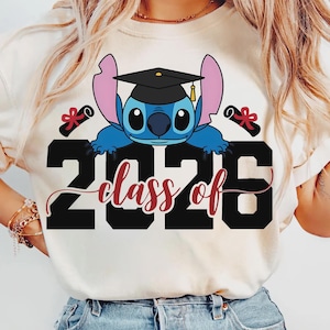 May include: Cream-colored t-shirt featuring a blue cartoon character wearing a graduation cap, with the text "2026 class of" in black and burgundy. Two graduation scrolls are on either side of the character.