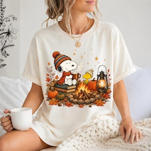 May include: A cream-colored short-sleeve t-shirt featuring a graphic of a cartoon dog wearing a knit hat and scarf, sitting on books by a campfire with a lantern, pumpkins, and autumn leaves. The design includes a small yellow bird character.