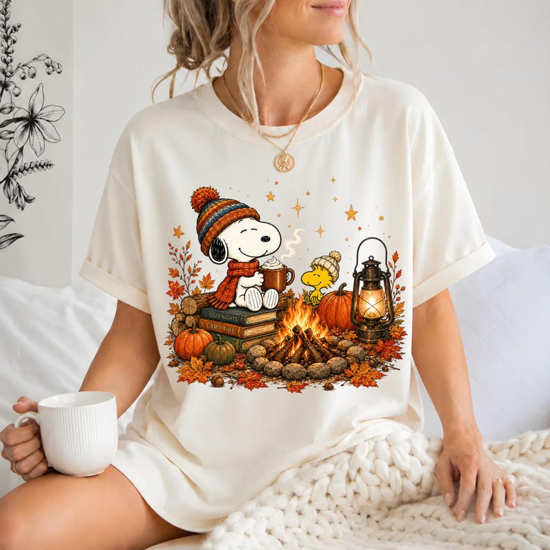 May include: A cream-colored short-sleeve t-shirt featuring a graphic of a cartoon dog wearing a knit hat and scarf, sitting on books by a campfire with a lantern, pumpkins, and autumn leaves. The design includes a small yellow bird character.