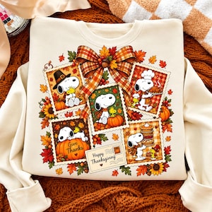Puede incluir: Una sudadera de cuello redondo color crema con un estampado gráfico de Snoopy y Woodstock en varias escenas de Acción de Gracias. El diseño incluye marcos estilo sello postal, hojas de otoño, girasoles y un gran lazo de cuadros arriba.