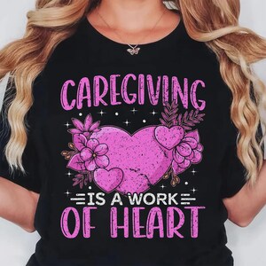May include: Black t-shirt with the words "Caregiving is a Work of Heart" in pink. The design features a pink heart with floral accents. The shirt is a crew neck.