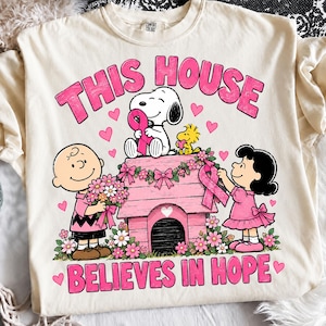May include: A cream-coloured t-shirt featuring a graphic of Snoopy, Charlie Brown, and Lucy with a pink doghouse. The text reads THIS HOUSE BELIEVES IN HOPE. The design includes pink breast cancer awareness ribbons, flowers, and hearts in shades of pink.
