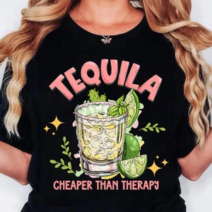 May include: Black t-shirt with a graphic of a tequila shot, lime, and mint. The word "TEQUILA" is in pink letters above the shot, and the phrase "CHEAPER THAN THERAPY" is below it. The design includes yellow stars and green leaves.