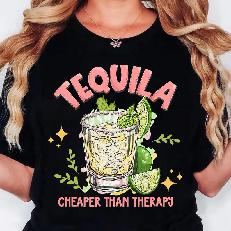 May include: Black t-shirt with a graphic of a tequila shot, lime, and mint. The word "TEQUILA" is in pink letters above the shot, and the phrase "CHEAPER THAN THERAPY" is below it. The design includes yellow stars and green leaves.