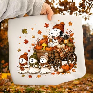 May include: A white fabric surface featuring a printed illustration of a white dog wearing a brown knit hat and scarf, sitting in a wooden wagon filled with pumpkins and a lantern. Three small white ghosts in knit hats pull the wagon.