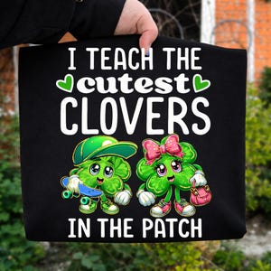 May include: Black sweatshirt with the text "I TEACH THE cutest CLOVERS IN THE PATCH". The graphic features two cartoon clovers, one with a skateboard and the other with a purse. The clovers are green with pink and white accents.
