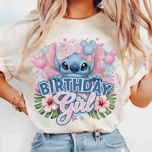 以下が含まれることがあります： クリーム色のTシャツ。青い漫画のキャラクターが「BIRTHDAY Girl」と書かれた看板を持っています。デザインにはピンクと青の風船、星、ハイビスカスの花が含まれています。キャラクターは大きな耳をしています。