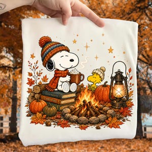 May include: A white t-shirt featuring a graphic of Snoopy wearing a colorful knit hat and scarf, holding a mug of cocoa while sitting on books labeled Cozy Nights and Camp Fire. Beside Snoopy is Woodstock, a pumpkin, and a lit lantern by a fire.