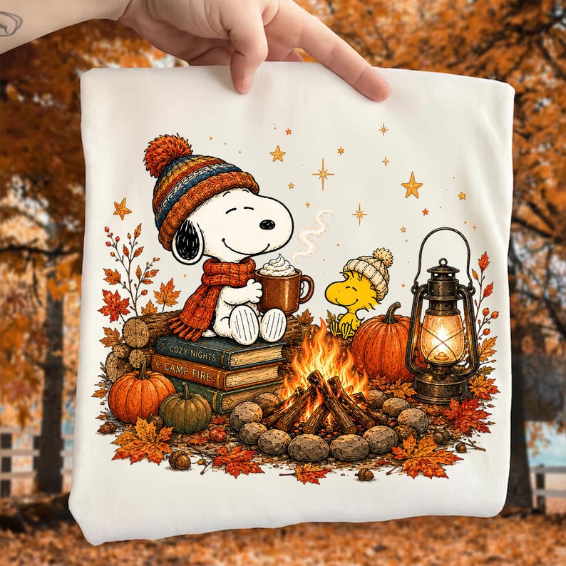 May include: A white t-shirt featuring a graphic of Snoopy wearing a colorful knit hat and scarf, holding a mug of cocoa while sitting on books labeled Cozy Nights and Camp Fire. Beside Snoopy is Woodstock, a pumpkin, and a lit lantern by a fire.