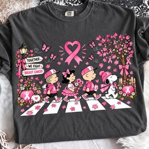 May include: A charcoal grey t-shirt featuring a graphic of four cartoon characters walking across a crosswalk. The design includes pink breast cancer awareness ribbons, butterflies, a tree, pumpkins, and a sign that reads Together We Fight Breast Cancer.