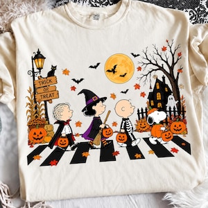 以下が含まれることがあります： ハロウィンの仮装をしたピーナッツのキャラクターたちが横断歩道を歩いているグラフィックが描かれたクリーム色のTシャツ。デザインには、満月、コウモリ、お化け屋敷、Trick or Treatと書かれた看板付きの街灯、カボチャが含まれています。