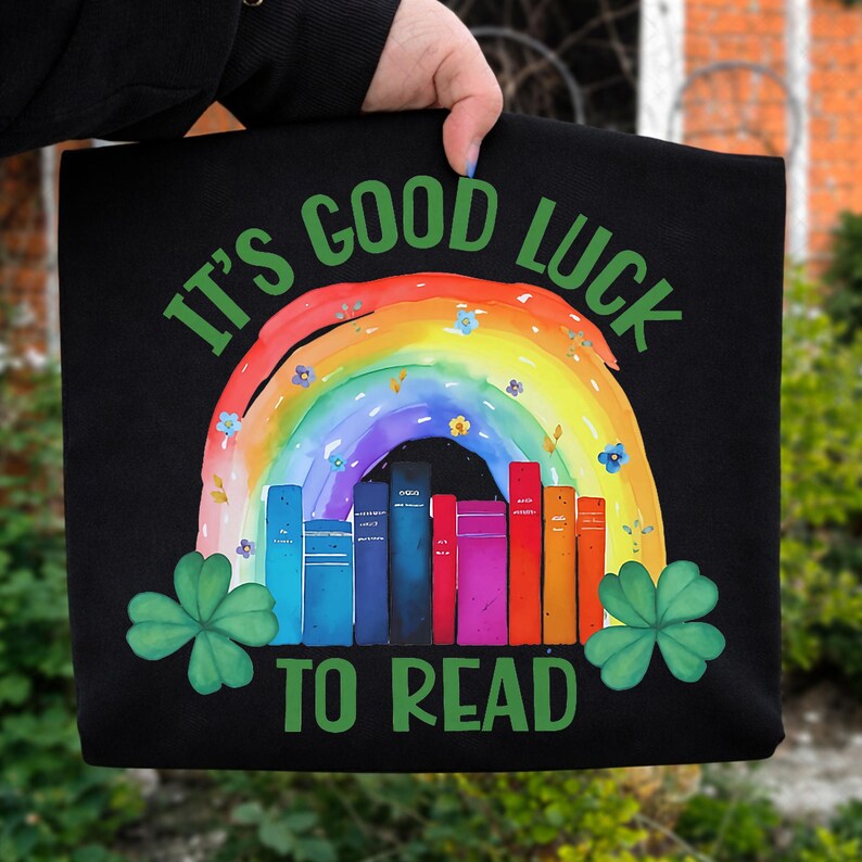 May include: Black tote bag with a rainbow and books design. The bag features the text "IT'S GOOD LUCK TO READ" in green. A rainbow arches over a row of colorful books, with shamrocks on either side.