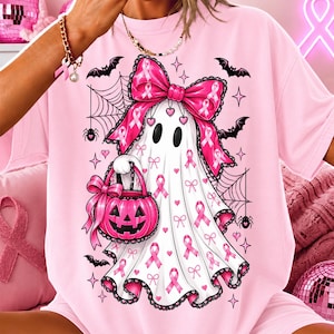 May include: A light pink t-shirt featuring a graphic of a white ghost wearing a large pink bow, holding a pink jack-o'-lantern bucket. The ghost is decorated with pink breast cancer awareness ribbons, surrounded by spiderwebs, bats, and stars.