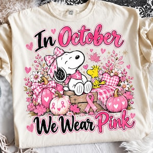 May include: A cream-coloured t-shirt featuring a graphic of Snoopy and Woodstock with pink pumpkins, flowers, and hearts. The text reads In October We Wear Pink. The design includes pink breast cancer awareness ribbons on the characters and pumpkins.