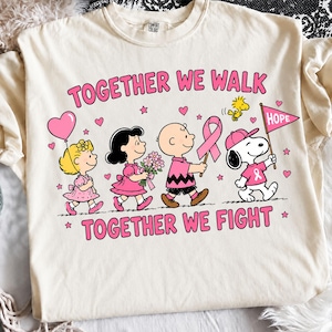 May include: A cream-coloured t-shirt featuring a graphic of Peanuts characters walking together. The text reads TOGETHER WE WALK and TOGETHER WE FIGHT. The design includes pink ribbons, hearts, stars, and a flag labelled HOPE in pink and black ink.