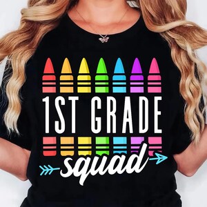 May include: Black t-shirt featuring a colorful crayon design above the words "1ST GRADE squad" in white. The design includes a row of crayons in red, orange, yellow, green, blue, purple, and pink. A fun back-to-school graphic tee.