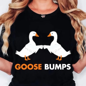 May include: Black t-shirt featuring a graphic of two white geese with orange beaks and feet, facing each other. The text "GOOSE BUMPS" is printed in orange below the geese. The shirt has a crew neck.