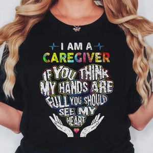 May include: Black t-shirt with the text "I AM A CAREGIVER" in colorful letters. The shirt features a heart-shaped design with the words "IF YOU THINK MY HANDS ARE FULL YOU SHOULD SEE MY HEART."