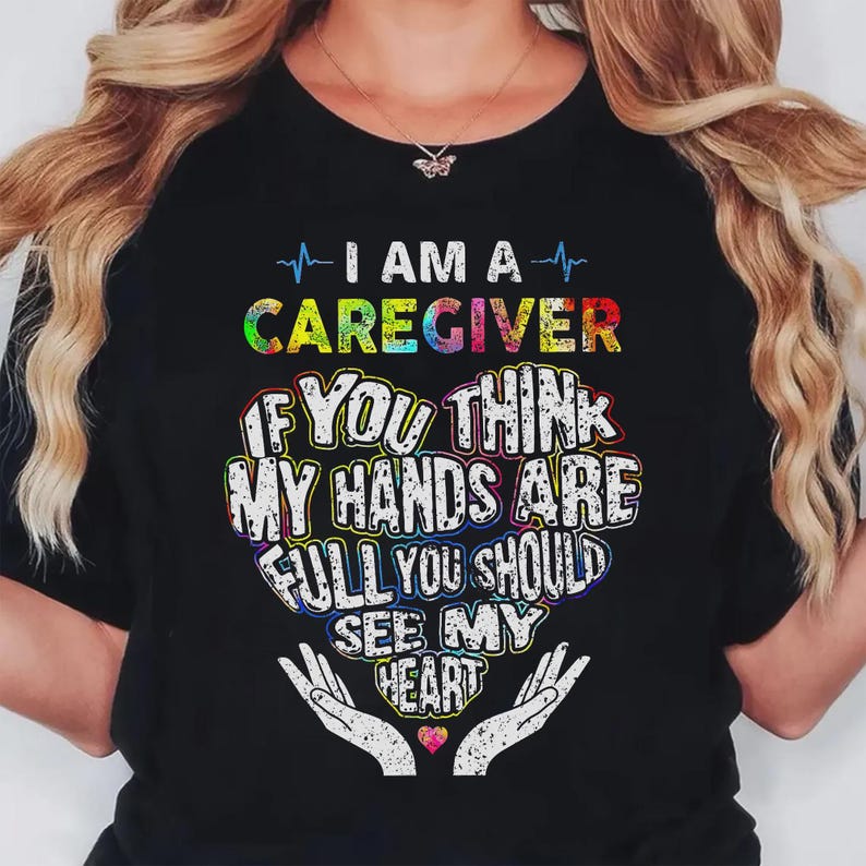 May include: Black t-shirt with the text "I AM A CAREGIVER" in colorful letters. The shirt features a heart-shaped design with the words "IF YOU THINK MY HANDS ARE FULL YOU SHOULD SEE MY HEART."